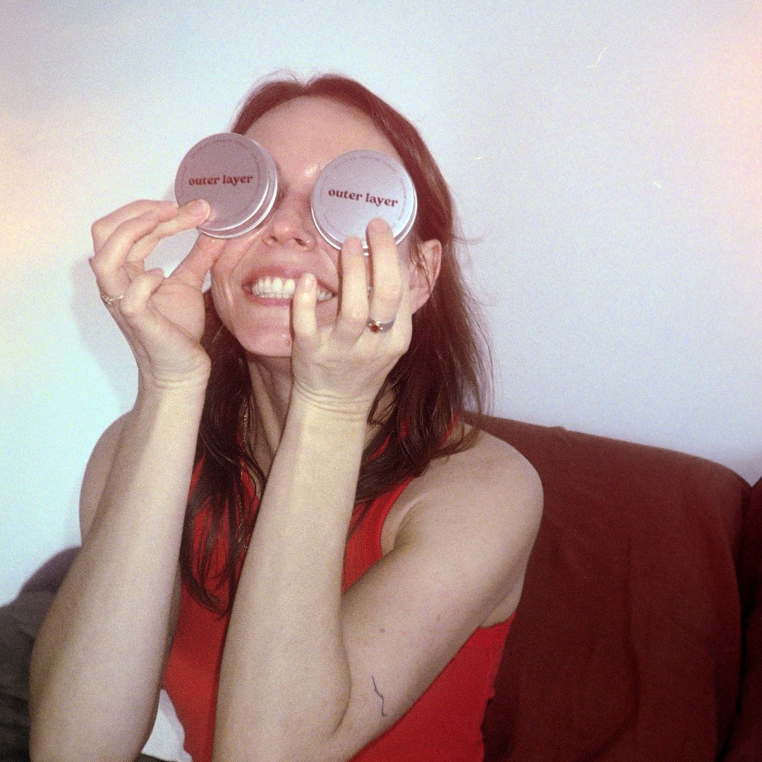 Smiling woman with brown hair holding two Outer Layer tins to her eyes against a white wall.